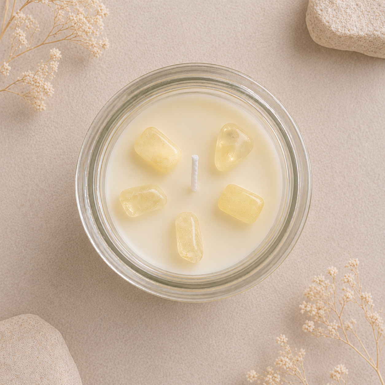 A top-down view of a glass jar candle with creamy off-white wax and five small pale yellow stone accents arranged in a circle around the wick. The candle sits on a neutral beige surface with dried flowers and stones as styled props.