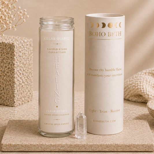 A clear glass jar candle labeled 'HEALING' from the BOHO BETH Sacred Stone Collection stands next to its matching cream-colored cylindrical packaging box on a stone surface, with a clear crystal pointed upward in front.