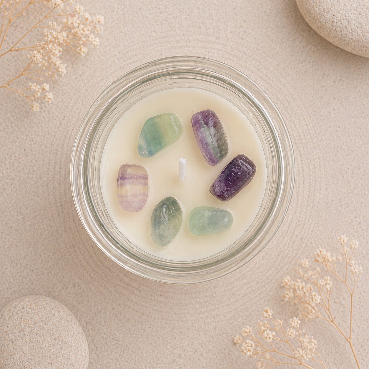 A clear glass jar candle filled with creamy off-white wax and five polished tumbled stones in green, purple, and lavender tones, arranged on a light beige textured surface with dried flowers and stone props.