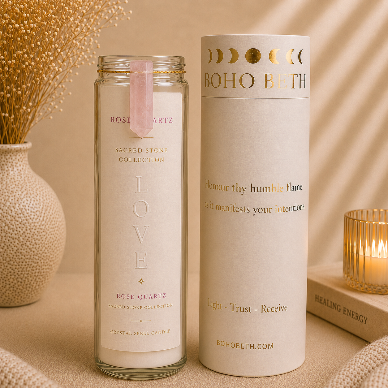 A glass jar candle and its matching cylindrical box from the Boho Bith Sacred Stones Collection, displayed on a beige surface with decorative dried grass, a lit candle, and a book in the background.