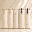 Five tall clear glass prayer candles with white wax are lined up on a light marble surface against a marble wall. Each candle has a different colored ribbon bookmark and is labeled with a sacred stone name: Citrine, Rose Quartz, Lapis Lazuli, Fluorite, and Amethyst.