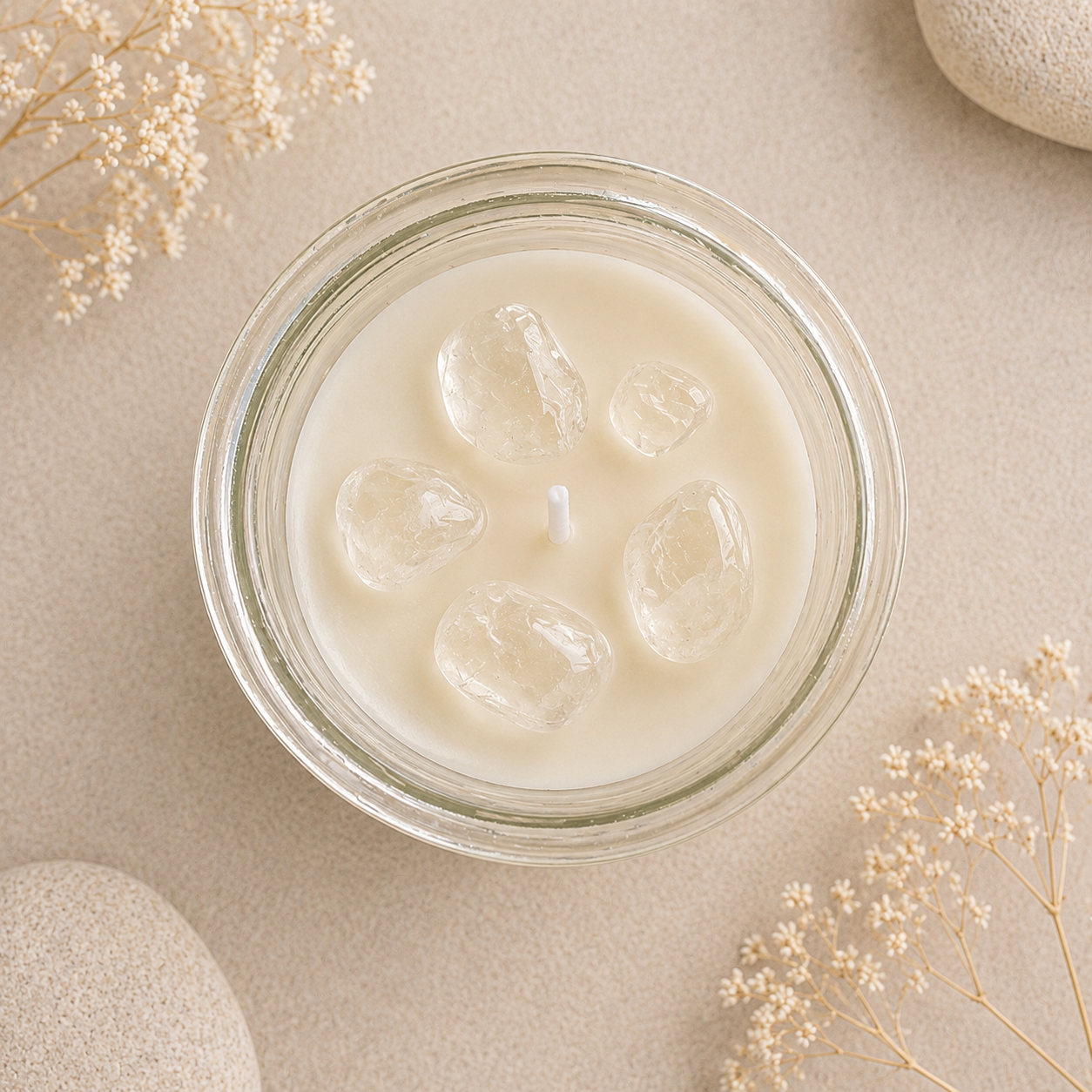Top-down view of a clear glass jar candle filled with creamy off-white wax and several clear ice cubes, styled on a beige surface with dried flowers and stone props.