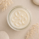 Top-down view of a clear glass jar candle filled with creamy off-white wax and several clear ice cubes, styled on a beige surface with dried flowers and stone props.
