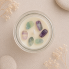 A clear glass jar candle filled with creamy off-white wax and five polished tumbled stones in green, purple, and lavender tones, arranged on a light beige textured surface with dried flowers and stone props.