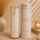 A glass jar candle and its matching cylindrical packaging box from Boho Beth's Sacred Stones Collection, featuring Citrine wax and gold lettering, styled on a beige fabric surface with dried flowers and a warm candle in the background.