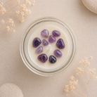 A clear glass jar candle filled with creamy off-white wax and decorated with smooth purple amethyst stones, photographed from above on a beige textured surface with dried flowers and stone props.
